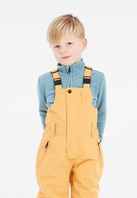 Gula vattentäta överdragsbyxor med justerbara axelremmar, tillsammans med en ljusblå fleece-tröja. Har sidofickor och en avslappnad passform.