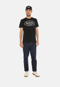 T-shirt en coton noir avec impression de logo crème, associé à un jean bleu foncé et des baskets blanches. Modèle portant une casquette noire assortie.