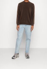 Sudadera marrón de cuello redondo, vaqueros azul claro y zapatillas grises. La sudadera tiene una textura suave; los vaqueros son ligeramente holgados.