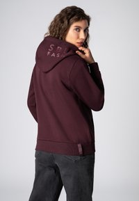 Bordeauxkleurige hoodie met een grote kap en geribbelde onderrand. Voorzien van "SBL" branding op de achterkant en textuurdetails in de stof.