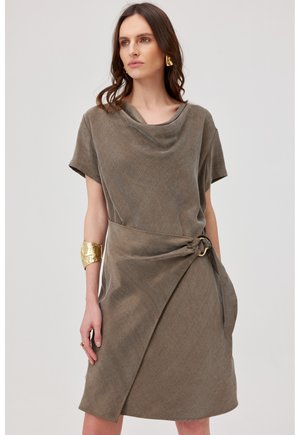 Kobieta ubrana w taupe sukienkę kopertową z krótkim rękawem, dekoltem z zakładkami, złotą bransoletką na mankiecie, złotymi kolczykami, stojąca na jednolitym tle.