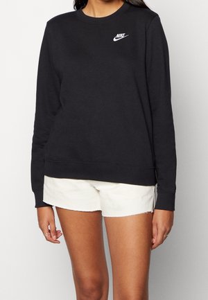 Kvinde iført en sort Nike langærmet sweatshirt og hvide shorts, vist fra nakke til midt på låret, stående mod en ensfarvet baggrund.