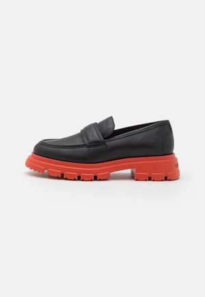 Mocassins en cuir noir avec une semelle en caoutchouc épaisse rouge, dotés d'un bout arrondi et d'un détail de sangle minimaliste sur le dessus.