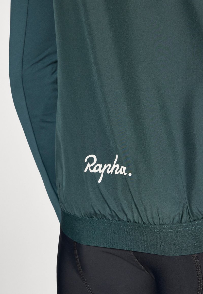 Veste de cyclisme vert foncé avec une texture douce, une taille élastiquée et le logo "Rapha." blanc sur le côté gauche. Elle possède des manches longues et un design ajusté.