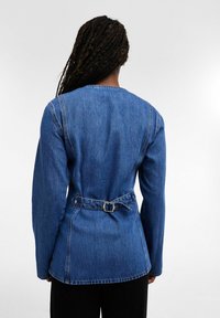 Femme aux longs cheveux tressés portant une veste en jean bleue cintrée avec une ceinture à la taille à l'arrière et un pantalon noir, vue de dos sur un fond blanc.