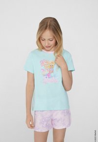 Světle modré tričko s krátkým rukávem a růžovým límcem, s graphic mermaidy a slovem "Barbie." V kombinaci s pastelově růžovými šortkami.