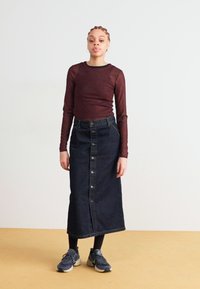 Μακριά σκούρα τζιν φούστα με κουμπιά μπροστά και πίσω σκίσιμο, συνδυασμένη με στενή μπορντώ μπλούζα με μακριά μανίκια. Το μοντέλο φοράει αθλητικά παπούτσια και κολάν.