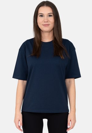 Giovane donna con capelli castani lisci che indossa una maglietta blu scuro tinta unita a maniche corte e pantaloni neri, in piedi davanti a uno sfondo bianco.