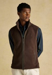 Hnedý fleecový vest s vysokým golierom a zipsom vpredu, s dvoma bočnými vreckami. Nosený cez modro-zelenú pruhovanú košeľu.