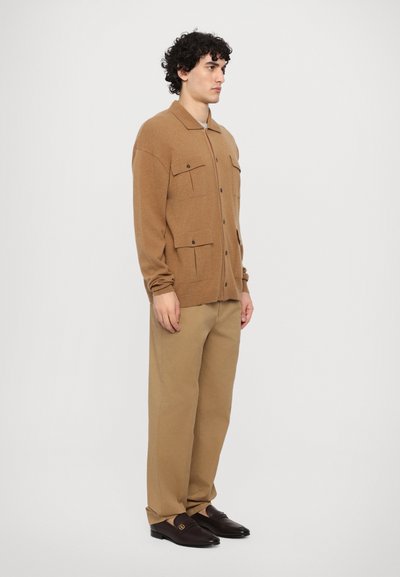 Cárdigan de punto marrón con cuatro bolsillos delanteros, cierre de botones y cuello con solapa, combinado con pantalones beiges y zapatos oscuros de piel sin cordones.