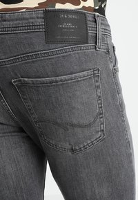 Szare dżinsy z denimu z tylną kieszenią ozdobioną detalami szwu oraz czarną, firmową metką na pasie. Gładka tekstura i prosty krój.