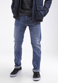 Jean en denim bleu avec une coupe décontractée, délavé clair, design à cinq poches et manches retroussées, associés à des baskets noires. Veste texturée bleu marine.