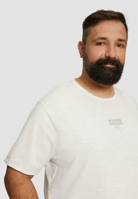 Biała koszulka z krótkim rękawem, wykonana z miękkiej tkaniny. Ma okrągły dekolt oraz małą metkę z logo na przodzie. Prosty design bez wzorów.