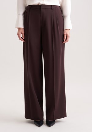 Pantalons évasés en tissu marron foncé, dotés d'une taille haute, de plis nets et d'une texture lisse, associés à des talons aiguille noirs.