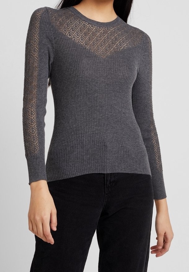 Femme portant un pull ajusté gris foncé à côtes avec des panneaux en dentelle en forme de losange sur les épaules et les manches, associé à un pantalon noir.