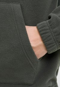 Dark green fleece hoodie featuring a front pocket, ribbed cuffs, and a soft texture. Visible hand partially inside the pocket.