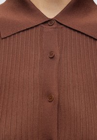 Chemise en maille côtelée marron avec un col pointu et trois boutons marron visibles fermés à l'avant.