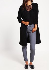 Cardigan negro de manga larga sobre una blusa de encaje, acompañado de jeans ajustados grises y zapatos planos negros. Tela suave y diseño en capas.