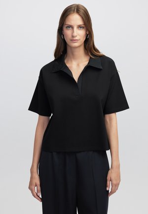 Polo shirt noir à manches courtes avec un col texturé, une patte de boutonnage à l'avant et une coupe ample. Le tissu semble lisse et doux.
