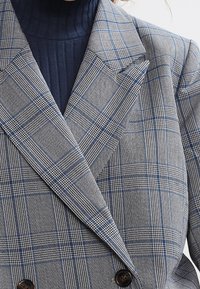 Harmaa ruudullinen kaksirivinen bleiseri, jossa on sinisiä ja valkoisia korostuksia, tekstuuripinta, huippulapelit ja suuret tummat napit.