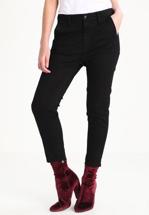 Person iført sorte smalle bukser og bordeaux fløjl ankle støvler, stående med den ene hånd i lommen mod en hvid baggrund.