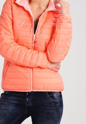 Personne portant une veste matelassée corail avec une doublure rose clair, zippée à mi-hauteur, assortie à un jean en denim bleu foncé sur un fond uni.