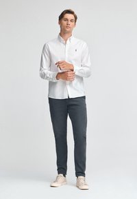 Camicia bianca con bottoni, maniche lunghe, in tessuto leggero, abbinata a pantaloni grigio scuro e sneakers chiare, indossata da un modello maschile in piedi.