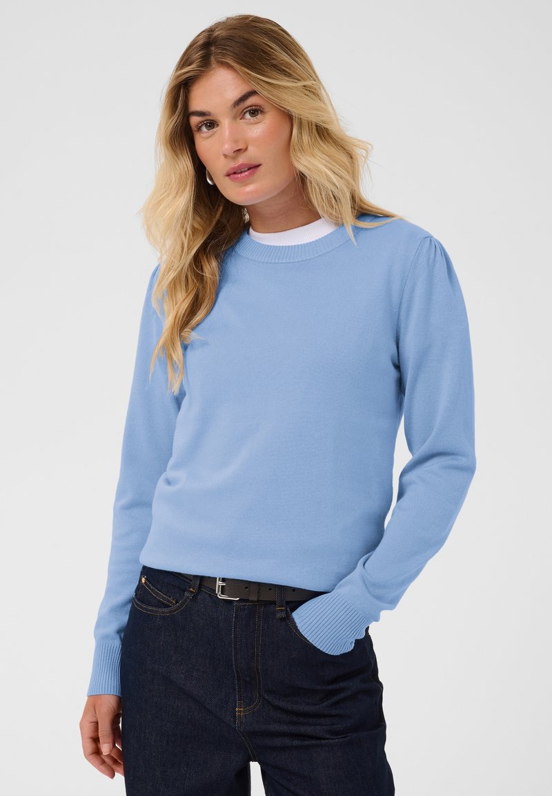 Femme blonde portant un pull bleu clair à manches longues, un jean foncé et une ceinture noire, posant avec une main dans la poche sur un fond blanc.
