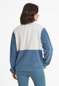 Woman facing away wearing a color-block fleece jacket with white upper and blue lower panels and matching blue leggings against a white background.