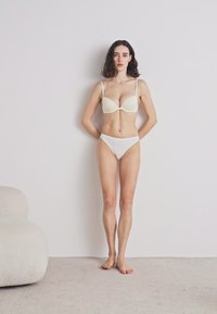 Beli set donjeg rublja s kalupljenim grudnjakom s tankim trakama i odgovarajućim bikinijem, prikazan na neutralnoj zidu i mekom tepihu.
