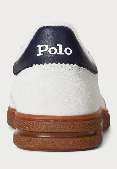 Baltas zomšinis sportbačių modelis su tamsiai mėlynu odiniu kulno akcentu, turintis tekstūruotą rudą gumos padą ir baltą "Polo" logotipą gale.