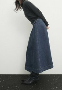 Denim-maksihame korkealla vyötäröllä, suora leikkaus, tummansininen väri ja sivuräystäs helman lähellä. Yhdistetty polveen asti ulottuviin mustiin saappaisiin.