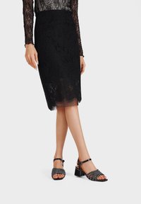 Falda lápiz de encaje negro que llega a la rodilla, combinada con sandalias de tacón negro con un diseño de cuentas curvas y correa en el tobillo.