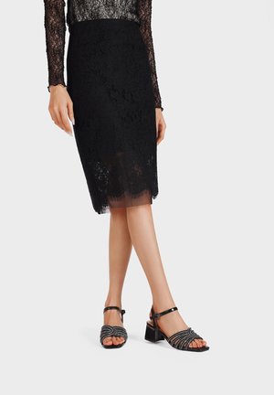 Gonna a matita in pizzo nero che arriva fino al ginocchio, abbinata a sandali neri con tacco caratterizzati da un design curvo con perline e cinturino alla caviglia.