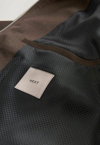 Veste marron avec une doublure intérieure noire à motifs et une étiquette carrée blanche cousue à l'intérieur portant l'inscription "NEXT".