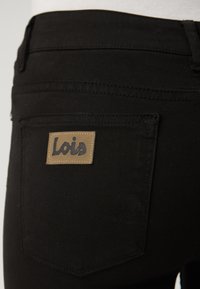 Jeans in denim nero con una tasca posteriore dotata di un'etichetta rettangolare color tabacco impresso con la scritta "Lois" in carattere nero. Texture liscia e design aderente.