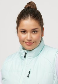 Gilet imbottito azzurro chiaro con collo alto e chiusura a zip, caratterizzato da una texture opaca e tiretti della zip neri. Dettaglio della tasca frontale visibile.