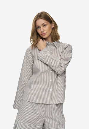 Femeie cu păr până la umeri poartă o cămașă cu dungi verticale bej și albe, cu nasturi, și pantaloni largi asortați, pe un fundal alb.