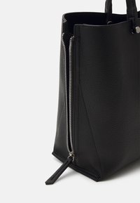Svart strukturert läder tote-väska med dragkedjestängning, vertikala sömdetaljer och korta handtag. Dragkedjehänget är en liten lädertag.