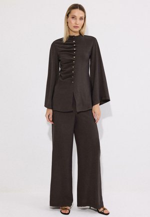 Γυναίκα με καφέ σκούρο μακρυμάνικο τοπ με κουμπιά μπροστά και αντίστοιχο φαρδύ παντελόνι, στέκεται με τα χέρια χαλαρά δίπλα στα πλευρά.