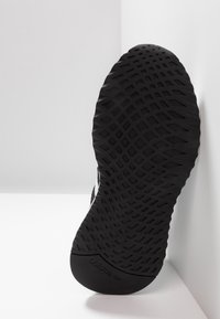 Svart sportskosula med ett strukturerat diamantmönster, med ett slätt hälområde och en inbäddad logotyp på undersidan.