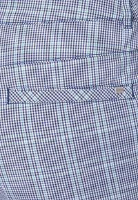 Sinivalkoinen ruudullinen kangas, jossa on ohuita turkooseja viivoja, sisältää takataskun ja hienovaraisen hopeisen logotagin helmassa.