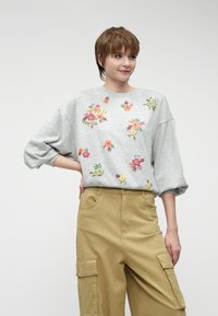 Sivá mikina s kvetinovou výšivkou v oranžovej, žltej a zelenej farbe, s voľnými rukávmi. Skombinovaná s khaki cargo nohavicami.