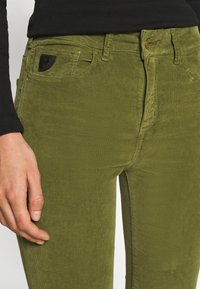 Pantalon en velours côtelé vert olive avec une coupe slim, présentant un détail de poche noire et des accessoires dorés à la taille. Texture douce avec des stries verticales.