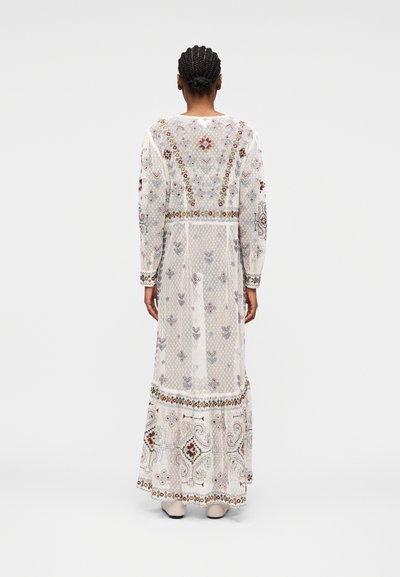 Femme aux cheveux tressés portant une longue robe blanche transparente ornée de broderies colorées florales et géométriques, debout devant un fond uni.