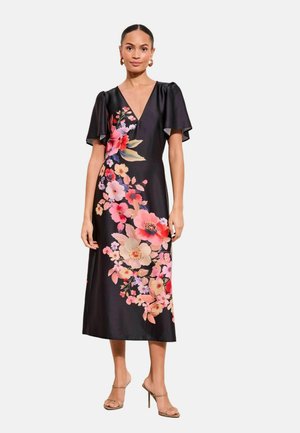 Kvinde iført en sort midi-kjole med flagrende ærmer og et diagonalt blomsterprint i nuancer af pink, rød og creme, kombineret med nøgenfarvede hæle.