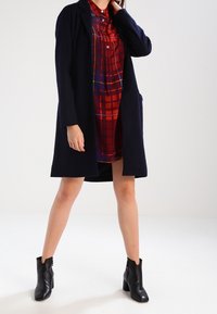Lungo cappotto blu navy sopra un vestito a quadri rossi, con colletto abbottonato, maniche lunghe e stivaletti alla caviglia con zip laterali. Tessuto dalla texture liscia.