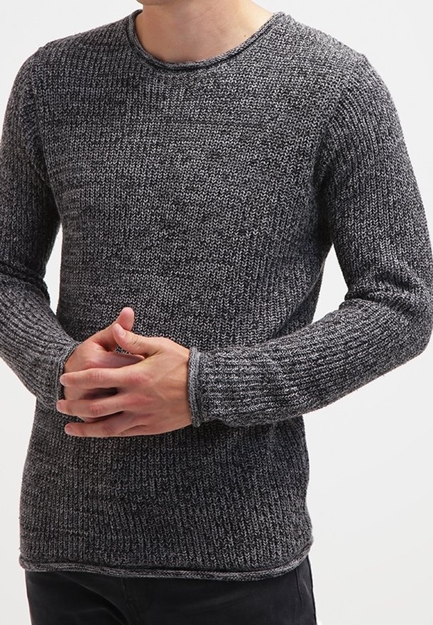 Person trägt einen strukturierten, dunkelgrauen Strickpullover mit langen Ärmeln, die Hände vor der unteren Brust gefaltet, vor einem schlichten Hintergrund.