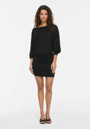 Une femme porte une robe noire aux manches amples et à la jupe ajustée, sur un fond de studio blanc uni.