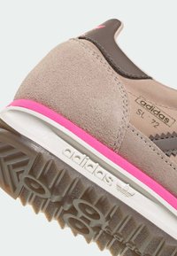 Hnědá semišová teniska s růžovými detaily, vybavená texturovanou podrážkou a výrazným logem. Klasický design, lehká konstrukce.